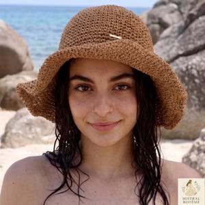 Peut inclure: Un chapeau de soleil marron tissé avec un large bord. Le chapeau a un petit accent de couleur or. Le chapeau est porté à l'extérieur près de l'océan. Le chapeau est un accessoire élégant pour la protection solaire.