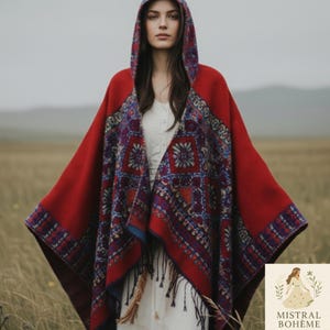 May include: A hooded red poncho with a patterned design in shades of blue, purple, and white. The poncho has a fringed hem and is worn over a white dress. The image is set in a field.