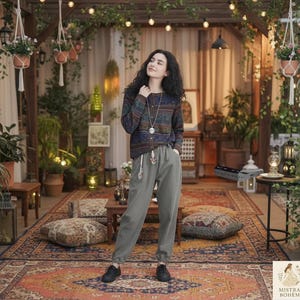 Puede incluir: Una mujer con un suéter estampado y pantalones verde oliva se encuentra en un ambiente de estilo bohemio. La escena incluye plantas colgantes, cojines decorativos y una alfombra vintage. La mujer lleva un collar largo y zapatos negros.