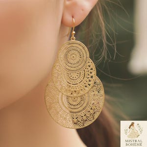 Peut inclure: Boucles d'oreilles pendantes dorées, composées de cercles ajourés superposés. Elles sont ornées de motifs complexes et sont présentées sur une oreille. Inspirées des styles bohème et ethnique.
