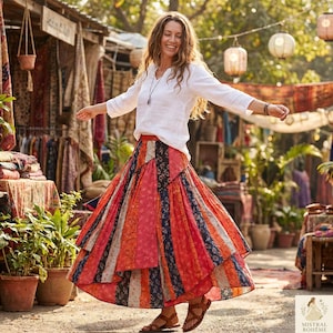 Peut inclure: Une femme porte une chemise blanche à col en V et une longue jupe à volants avec des rayures verticales rouges, oranges et marine. La jupe est de style bohème. La femme sourit et porte des sandales. L'arrière-plan montre un marché.