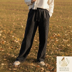 Peut inclure: Pantalon noir à jambes larges avec un motif subtil de points bleus. Le pantalon a une taille élastique avec cordon de serrage et des poches. Le modèle porte des baskets blanches et un pull blanc. Collection Mistral Bohème.