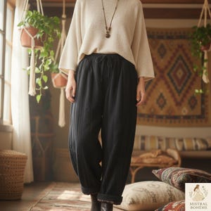 Peut inclure: Une personne porte un pull crème et un pantalon noir matelassé avec une taille élastique. Le pantalon est retroussé aux chevilles. Un long collier avec un pendentif est visible. L'image est prise dans une pièce avec des plantes suspendues et un tapis à motifs.