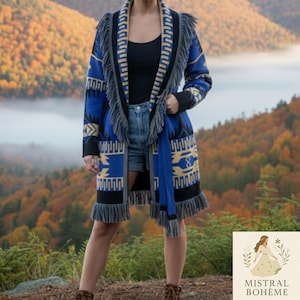 Op de afbeelding: Lange, blauw, zwart en beige patroon cardigan met franjes. De cardigan heeft een geometrisch ontwerp en wordt gedragen over een zwarte top en een spijkerbroek. De achtergrond toont een schilderachtig uitzicht op een bergketen.