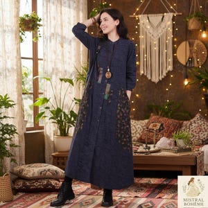 Könnte beinhalten: Ein marineblaues Langarmkleid mit Knopfleiste, dekorativen Patches und floralen Mustern. Das Kleid hat einen Stehkragen und Taschen. Das Model trägt schwarze Stiefel und eine lange Halskette. Das Markenlogo "MISTRAL BOHEME" ist sichtbar.