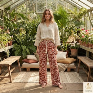 Peut inclure: Une femme porte un haut en lin beige et un pantalon palazzo à motifs avec une taille à cordon. Le pantalon présente un motif répétitif dans les tons de rouge et de crème. Le cadre est une serre avec une verdure luxuriante et des bancs en bois.