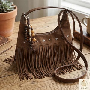 Peut inclure: Sac à bandoulière en daim marron avec franges et longue sangle. Le sac est orné d'étoiles et d'un charm en perles. Il est posé sur une surface en bois.