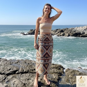 Peut inclure: Un haut tube beige et une longue jupe à motifs avec des motifs géométriques dans les tons marron, bleu et jaune. La jupe a une taille haute et une silhouette droite. La personne se tient sur des rochers près de la mer.