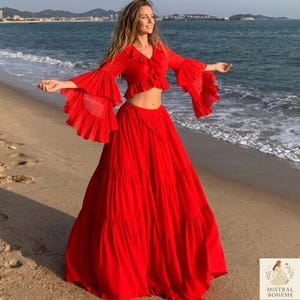Op de afbeelding: Een felrode tweedelige outfit op een strand. De top heeft een gerimpelde halslijn en uitlopende mouwen. De lange rok is gelaagd. De oceaan en de lucht zijn op de achtergrond. Het Mistral Bohème-logo staat rechtsonder.