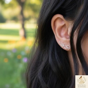 Peut inclure: Boucle d'oreille grimpante argentée avec un design délicat et tourbillonnant. La boucle d'oreille épouse l'oreille, présentant un motif complexe de boucles et de volutes. Le bijou est présenté sur une oreille, avec un arrière-plan flou de verdure.