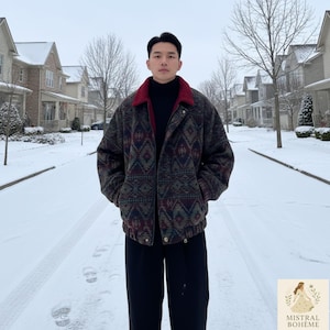 Op de afbeelding: Een jas met patroon met een rode kraag, een zwarte broek en een zwarte coltrui. De jas heeft een geometrisch ontwerp in tinten bruin, groen en bordeaux. De persoon staat in een besneeuwde omgeving.