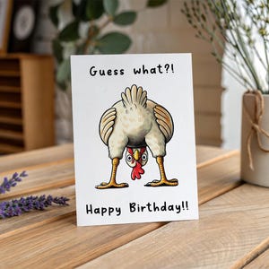 May include: A white greeting card with a cartoon chicken illustration. The chicken is upside down, with its rear end facing the viewer. The text "Guess what?!" and "Happy Birthday!!" are printed on the card.