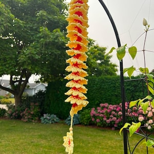 May include: A hanging spiral decoration crafted from yellow and red yarn, featuring a series of ruffled, flower-like crochet layers arranged in a vertical column, finished with a single dangling flower at the bottom and a small wooden bead.
