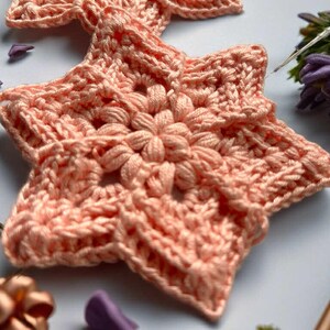 May include: Close-up of two peach-colored, handmade crochet star ornaments. Each star features a central flower design and detailed stitching. The stars are placed on a light blue surface, with decorative elements in the background.