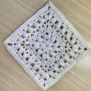 Puede incluir: Un tapete cuadrado de ganchillo blanco roto con un diseño floral en el centro. La pieza hecha a mano presenta un patrón texturizado y en relieve con un borde calado detallado. El tapete probablemente está hecho de algodón o un hilo similar.
