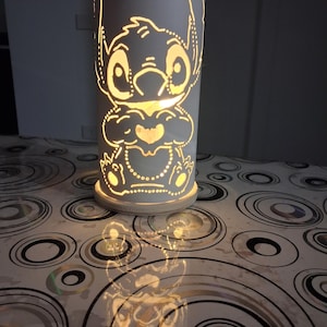 Può includere: Lampada cilindrica bianca con un design intagliato del personaggio Stitch. La lampada emette una luce gialla calda, evidenziando i tratti di Stitch. La lampada è posizionata su una superficie con un motivo circolare. La lampada è alta circa 20 cm.