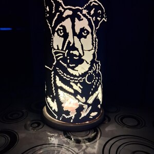 Può includere: Lampada cilindrica nera con silhouette di cane bianca. La lampada è illuminata dall'interno, creando un bagliore caldo. Il design del cane include un collare. La lampada poggia su una base circolare.