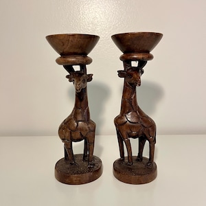 May include: Two carved wooden giraffe candle holders. The giraffes are brown with detailed carvings, supporting a bowl-shaped top. The candle holders are on a white surface.