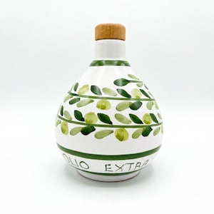 May include: A white ceramic oil cruet with a rounded body and a light brown wooden stopper. The surface features hand-painted green olive branches and leaves arranged in horizontal bands, with the text OLIO EXTRA printed in black lettering.