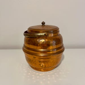 May include: A hammered copper container with a lid and a handle. The container has a round shape with horizontal bands and a small knob on the lid. The handle is a brass color. The copper has a warm, reddish-brown hue.