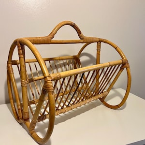 May include: A vintage, light brown rattan magazine rack. The rack features a handle and curved sides, with a slatted interior for holding magazines or newspapers. The piece is made of woven rattan.