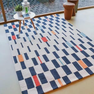 May include: A rectangular rug featuring a geometric design of navy blue rectangles on a cream backdrop. It has orange, red, and light blue accents. The rug is placed on a light-coloured floor.