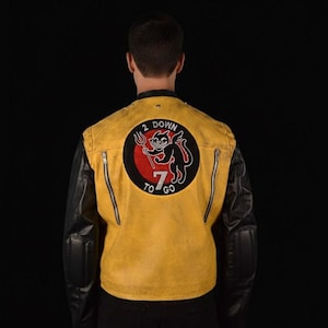 Può includere: Giacca in pelle gialla e nera con grafica di diavolo sul retro. La giacca ha maniche nere e corpo giallo con due cerniere verticali. La grafica recita "2 Down 7 to Go".