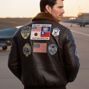 Puede incluir: Chaqueta bomber de cuero marrón oscuro con múltiples parches, incluyendo la bandera estadounidense, una bandera japonesa y el texto "FAR EAST CRUISE 63-4" y "USS GALVESTON". La chaqueta tiene un cuello de piel marrón y se lleva sobre una camisa azul.