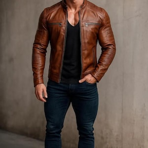 May include: A brown leather jacket with a stand-up collar and zipped pockets. The jacket features quilted detailing on the shoulders and sleeves. The person in the image is wearing a black v-neck shirt and dark blue jeans.