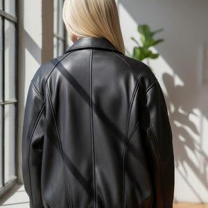 Puede incluir: Chaqueta negra de cuero sint&eacute;tico con dise&ntilde;o tipo bomber. La chaqueta presenta dobladillo y pu&ntilde;os acanalados, cuello y detalles de costuras verticales. La parte trasera de la chaqueta es visible, mostrando el ajuste a medida y la apariencia elegante.