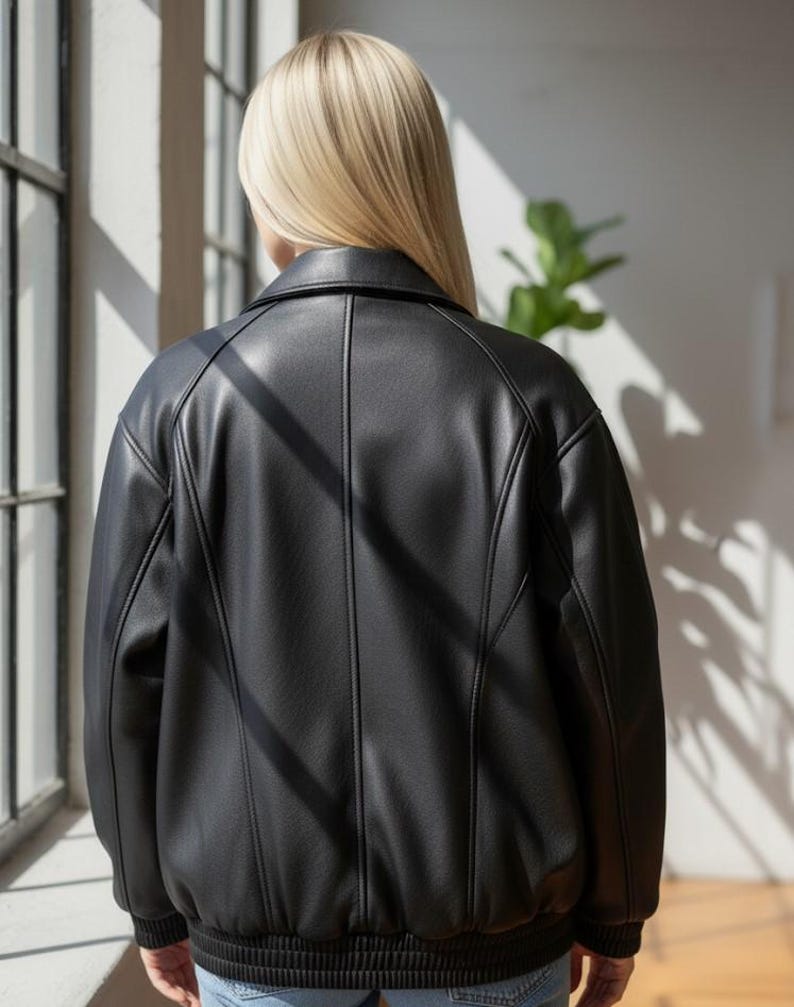 Puede incluir: Chaqueta negra de cuero sint&eacute;tico con dise&ntilde;o tipo bomber. La chaqueta presenta dobladillo y pu&ntilde;os acanalados, cuello y detalles de costuras verticales. La parte trasera de la chaqueta es visible, mostrando el ajuste a medida y la apariencia elegante.