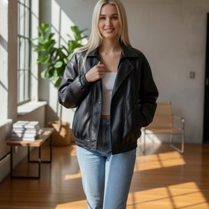 Puede incluir: Chaqueta de cuero negra con cuello y cierre de botones. La chaqueta tiene dobladillo y pu&ntilde;os acanalados. La persona lleva una camiseta blanca y vaqueros azul claro. El fondo incluye una ventana, una planta y un banco de madera.