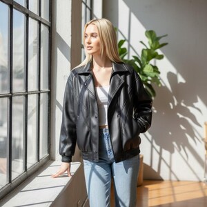 Puede incluir: Chaqueta bomber de cuero sint&eacute;tico negro con cuello con muescas, cierre de botones y pu&ntilde;os acanalados. La chaqueta se combina con vaqueros azul claro. El modelo est&aacute; de pie cerca de una ventana grande.