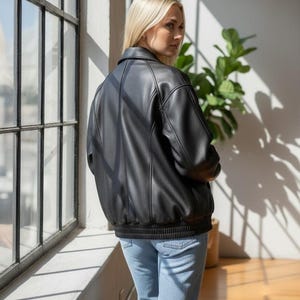 Puede incluir: Chaqueta bomber negra con dobladillo y pu&ntilde;os de canal&eacute;. La chaqueta tiene un cuello cl&aacute;sico y detalles de costura verticales. La modelo lleva vaqueros azul claro. El fondo incluye una ventana y una planta en maceta.