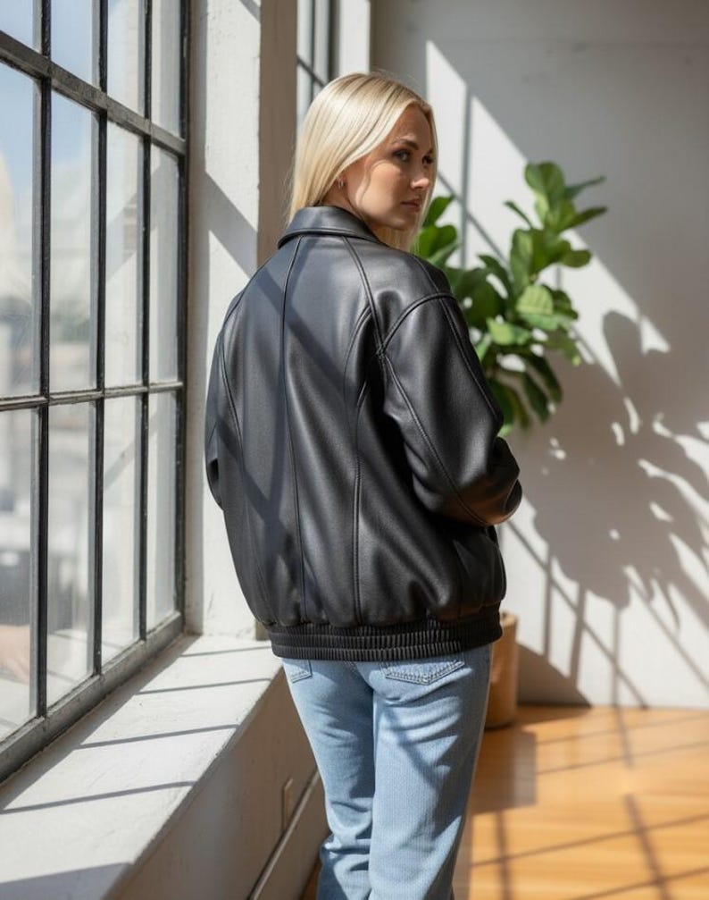 Puede incluir: Chaqueta bomber negra con dobladillo y pu&ntilde;os de canal&eacute;. La chaqueta tiene un cuello cl&aacute;sico y detalles de costura verticales. La modelo lleva vaqueros azul claro. El fondo incluye una ventana y una planta en maceta.