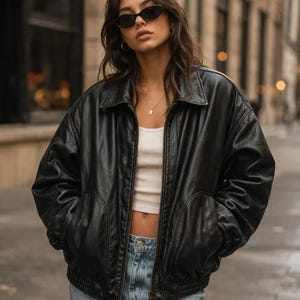 Puede incluir: Chaqueta bomber de cuero negro con cuello y bolsillos delanteros. La chaqueta se lleva sobre un top corto blanco y vaqueros azules. La persona lleva gafas de sol negras y un collar.