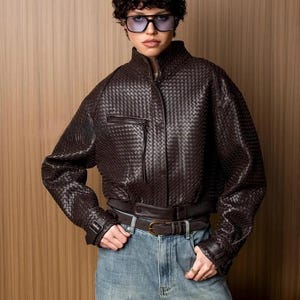 Può includere: Giacca in pelle intrecciata marrone scuro con collo alto e cerniera frontale. La giacca ha una piccola tasca sul petto ed è abbinata a jeans blu e una cintura marrone. La modella indossa occhiali da sole scuri.
