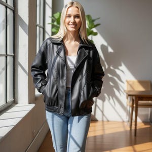 Puede incluir: Chaqueta negra de cuero sint&eacute;tico con cierre de botones y pu&ntilde;os acanalados. La chaqueta tiene un estilo bomber cl&aacute;sico con cuello. La persona lleva vaqueros azul claro y una camiseta blanca. El fondo incluye una ventana y una planta.