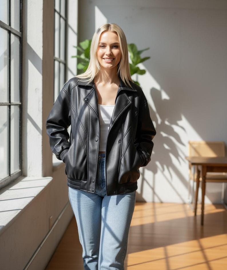 Puede incluir: Chaqueta negra de cuero sint&eacute;tico con cierre de botones y pu&ntilde;os acanalados. La chaqueta tiene un estilo bomber cl&aacute;sico con cuello. La persona lleva vaqueros azul claro y una camiseta blanca. El fondo incluye una ventana y una planta.