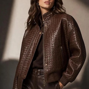 May include: A dark brown bomber jacket featuring a woven leather texture, a high collar, a front zipper closure with snap buttons, and ribbed cuffs. The jacket is worn over a dark brown sweater and paired with dark brown leather trousers.
