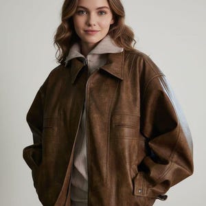May include: A brown leather jacket with a collared neckline, a front zipper closure, and two horizontal slit pockets on the chest. The jacket features long sleeves with cuffs and an elasticized hem, worn over a beige high-neck sweater with a zipper.