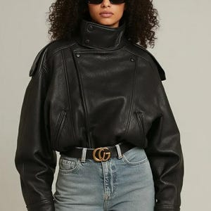Puede incluir: Chaqueta de cuero negra con cuello alto y cierre cruzado. La chaqueta se combina con vaqueros azul claro y un cinturón negro con hebilla dorada. La persona lleva gafas de sol negras.