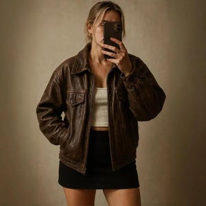 May include: A brown leather jacket with a collared neckline and two front pockets. The jacket is worn over a white tank top and a black mini skirt. The person in the image is holding a phone in front of their face.