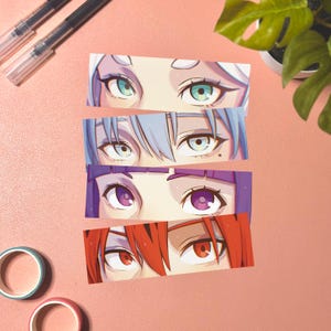Puede incluir: Cuatro pegatinas rectangulares con ojos de estilo anime con iris y cabello de diferentes colores. Los ojos son de color verde, azul, morado y rojo. Las pegatinas están dispuestas sobre una superficie rosa con bolígrafos, cinta washi y una planta en el fondo.