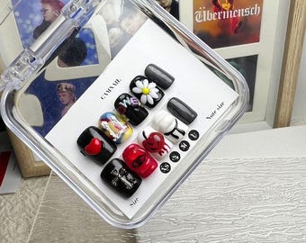 May include: A set of ten press-on nails in a clear plastic case, featuring various designs including a white daisy on black, a red heart, a silver cross, a red cherry, and colourful abstract patterns. The nails are arranged on a white card.