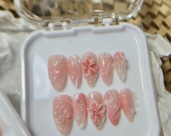 Może przedstawiać: Zestaw sztucznych paznokci w przezroczystym plastikowym etui. Paznokcie są głównie różowe z białymi akcentami i kwiatowymi wzorami. Niektóre paznokcie mają trójwymiarowe ozdoby kwiatowe i małe dżety. Paznokcie są ułożone w rzędzie.