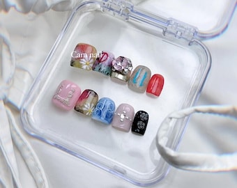 May include: A collection of ten false nails in a clear plastic case. The nails display diverse designs, such as floral patterns, rhinestones, and abstract art. The text "Cam nail" is visible on one nail.