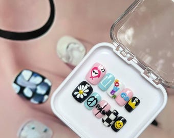 May include: A collection of press-on nails featuring diverse designs such as a daisy, peace symbol, and a smiley face. The nails are presented in a white plastic case with a transparent lid. The backdrop is a soft pink hue.