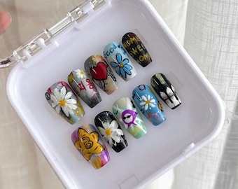 May include: A collection of ten false nails, each decorated with individual designs. The designs include daisies, hearts, and text, presented in a white plastic case. The nails feature a variety of colours, including blue, yellow, and white.