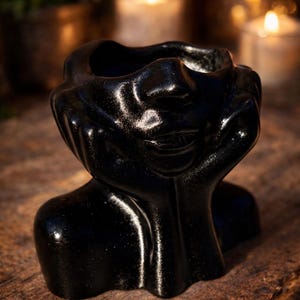 Könnte beinhalten: Ein schwarzer, glitzernder Übertopf in Form eines menschlichen Kopfes, der auf zwei Händen ruht. Die Skulptur hat eine glatte, reflektierende Oberfläche mit feinen Glitzerpartikeln im dunklen Finish und bildet oben eine hohle Gefäßöffnung.